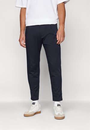 Homme portant un pantalon court bleu marine, des chaussettes blanches, des baskets blanches et une chemise blanche à manches courtes sur un fond uni.