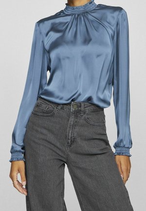 Kvinde iført en skinnende blå langærmet bluse med smockkrave og manchetter, kombineret med højtaljede sorte denimjeans.