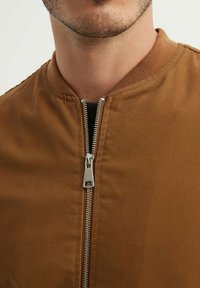 Veste bombardier marron avec fermeture éclair, col côtelé et texture de tissu durable. Gros plan mettant en évidence les coutures et les détails du matériau.