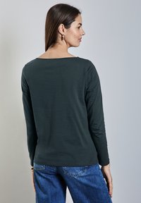 Tummanvihreä pitkähihainen paita, jossa on leveä kaula-aukko ja sileä pinta, yhdistettynä sinisiin farkkuihin, joissa on klassinen leikkaus ja takataskut.