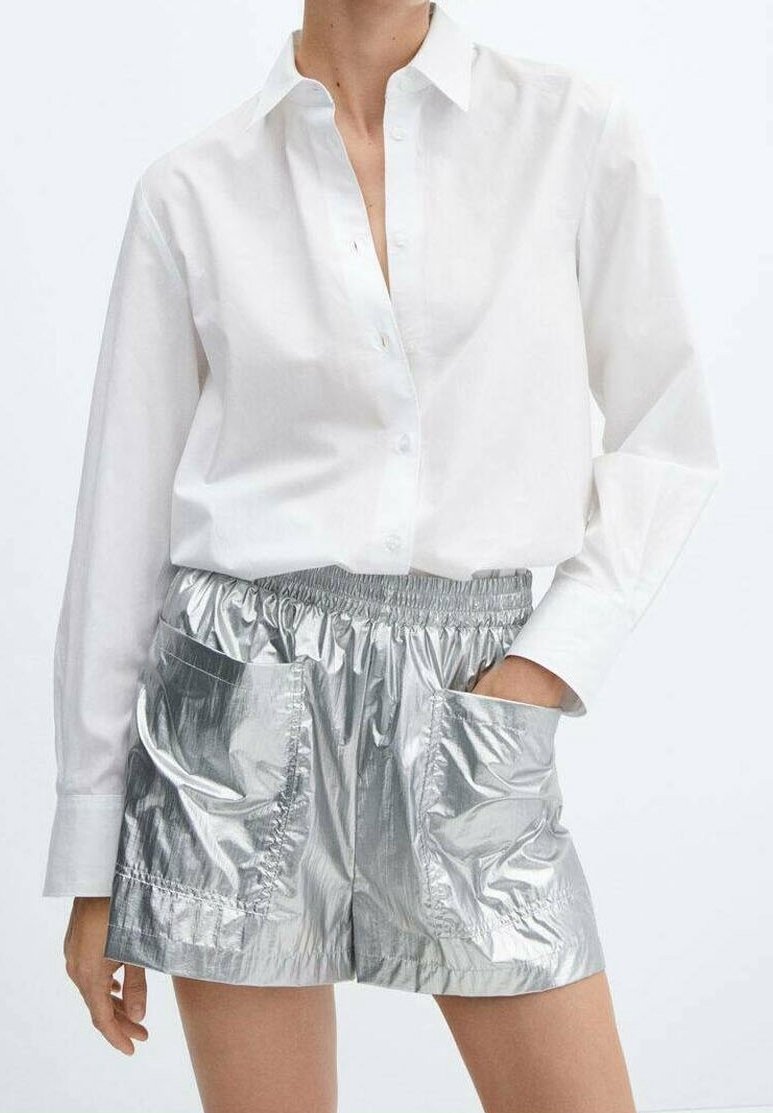 La personne porte une chemise blanche à manches longues boutonnée, rentrée dans un short argenté brillant à taille haute, avec de grandes poches avant et une ceinture élastique.