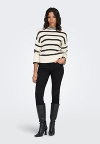 Roomwit-zwart gestreepte trui met een hoge halslijn, gecombineerd met zwarte skinny jeans en knielange zwarte laarzen. Soepele textuur en getailleerd ontwerp.