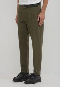 Olivgrüne, plissierte Hosen aus einem Baumwollmischgewebe, mit geradem Bein, seitlichen Taschen und kombiniert mit schwarzen Schuhen.