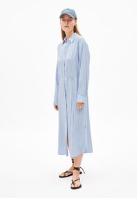 Robe longue à rayures bleu et blanc avec un devant boutonné, un col et des manches longues ; associée à des sandales plates noires à lanières.