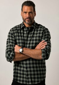 Camicia di flanella a quadri verdi e neri con maniche lunghe, colletto con bottoni, modello che indossa un orologio, braccia incrociate su uno sfondo neutro.