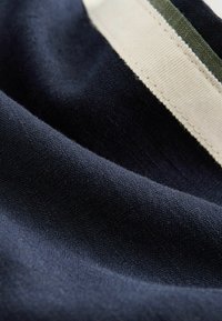 Granatowa bawełniana tkanina o gładkiej fakturze, z kontrastującymi paskami w kolorze khaki i kremowym wzdłuż jednej krawędzi. Zbliżenie podkreśla szczegóły.