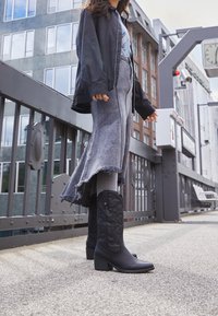 Černé kožené kotníkové boty s texturovaným vzorem a blokovými podpatky, doplněné dlouhou šedou džínovou sukní s třepeným lemem.