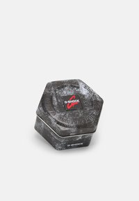 Boîte en métal hexagonale G-SHOCK avec une finition noir mat et une surface texturée, ornée d'un logo rouge sur le couvercle. Des accents incluent du texte en relief.