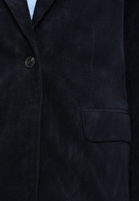 Mornarsko plavi blazer od kordurovog materijala s urezanim reverom, jednim gumbom za zatvaranje i zakrpnim džepom na lijevoj strani. Vidljiv je detalj teksturirane tkanine.