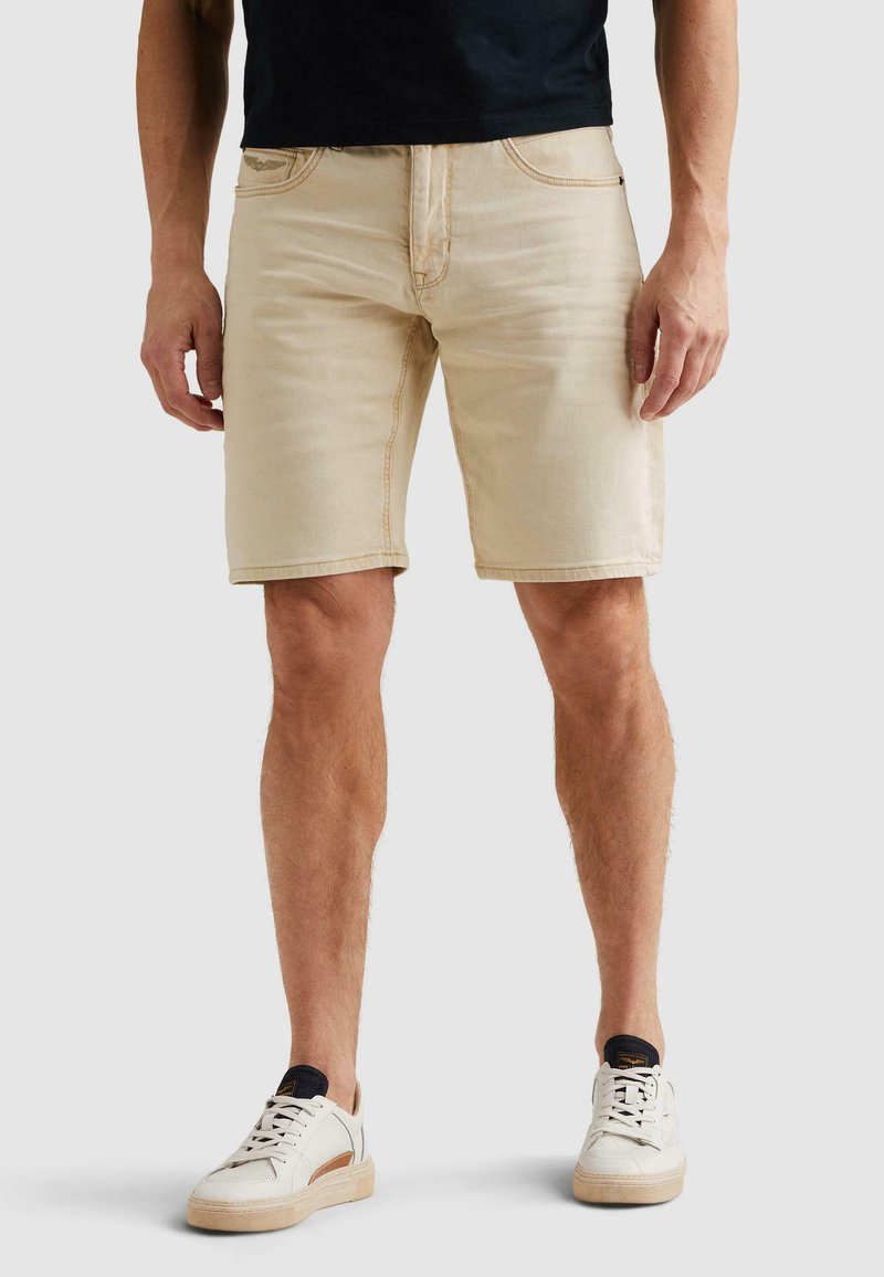 Homme portant un short en jean beige, une chemise noire et des baskets blanches, debout devant un fond uni.