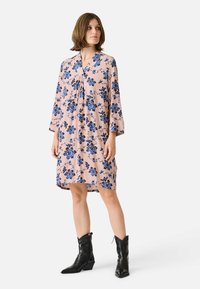 Rochie florală de culoare roz deschis, cu flori albastre, cu mâneci lungi, decolteu în V și croială lejera. Combinată cu botine negre până la gleznă.