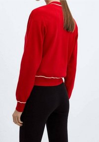 Frau mit langen, glatten Haaren trägt einen roten Pullover mit weißen Besätzen an den Ärmeln und schwarze, eng anliegende Hosen, von hinten gezeigt.