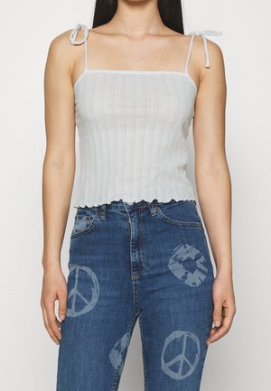 Vrouw draagt een wit geribd mouwloos topje met strikbandjes en een blauwe spijkerbroek met gebleekte vredestekenpatronen.