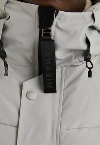 Gros plan sur une veste gris clair avec une tirette de fermeture éclair noire et des cordons de serrage, présentant un bouton-pression en métal gravé du mot "CHASIN".