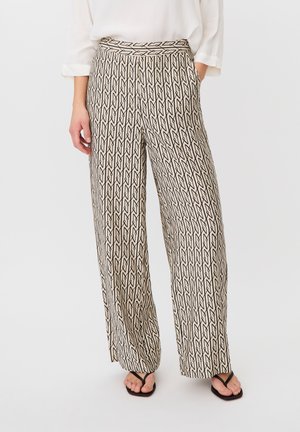 Person iført beige bukser med brede ben og sort geometrisk mønster, hvid bluse og sorte sandaler, stående mod en ensfarvet baggrund.