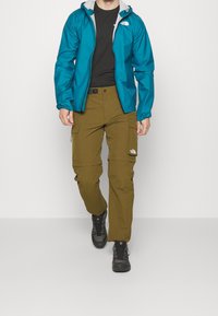 Niebieska wodoodporna kurtka z szarym kapturem, czarna koszulka oraz oliwkowe spodnie cargo z kieszeniami i zamkami, w połączeniu z czarnymi butami trekkingowymi.