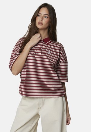 Jeune femme aux longs cheveux bruns, vêtue d'un polo rayé rouge et blanc et d'un pantalon ample beige, posant avec une main près du cou.