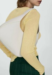 Sac à main en cuir blanc à la forme sculpturale. La bandoulière est large et la texture semble lisse. Le modèle porte un haut côtelé jaune et une jupe verte.