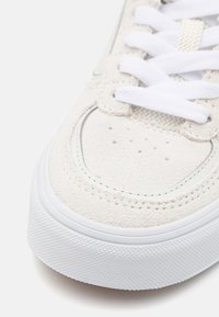 Zapatilla de ante beige claro con cordones blancos y suela de goma. Presenta perforaciones en la puntera y borde blanco en los laterales.