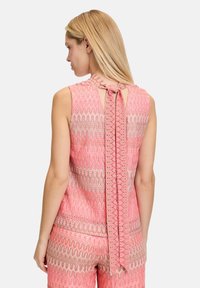 Ärmelloses, pinkfarbenes Oberteil mit Muster, einem Ausschnitt im Rücken und einer Schleifenverschlüsse. Das strukturierte Design zeigt wellenförmige Linien in unterschiedlichen Schattierungen von Pink und Beige.