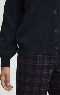 Gilet en tricot bleu marine avec une finition texturée, présentant de grands boutons et des manches bouffantes, associé à un pantalon à carreaux noir et rose.