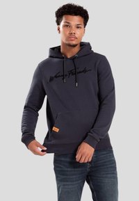 Donkerblauw hoodie met een kangoeroezak, zwart geborduurd logo, verstelbare koorden en een oranje accentlabel aan de rand van de zak.