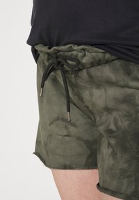 Calções verde-oliva feitos de um tecido suave, com um cós ajustável com cordão e pontas prateadas, e uma bainha desfiada. Textura de padrão subtil.
