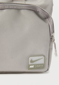 Γκρι υφασμάτινη τσάντα Nike Utility 2.0 γωνία με φερμουάρ και ραμμένη ετικέτα λογότυπου με το σήμα Nike και κείμενο.