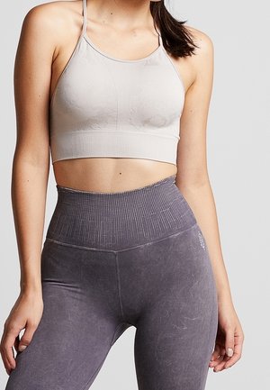 Nainen, joka käyttää vaaleanharmaata urheiluliivejä ja korkeavyötäröisiä, tekstuurillisia tummanharmaita leggingssejä, seisoo yksinkertaisen taustan edessä.