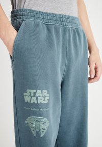 Helle türkisfarbene Jogginghosen aus weichem Stoff, mit einem gedruckten "Star Wars"-Logo und einer Grafik des Millennium Falcon mit Textdetails.