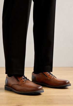 Brązowe skórzane buty wizytowe z perforowanym wzorem, czarnymi akcentami i niebieskimi sznurówkami, w zestawieniu z czarnymi dopasowanymi spodniami.