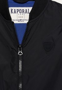 Musta pommitakki, jossa on vetoketjukiinni, pehmeä kangas, neidotettukraavi ja kohotettu logopatch. Sisäpuolella on sininen verkkomateriaali.