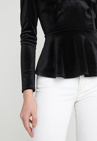 Musta pitkähihaine samettinen peplum-toppi, joka on yhdistetty valkoisiin kapeisiin farkkuihin, näyttäen vartalon olkapäästä yläreiteen.