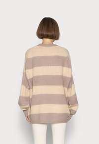 Person med kort brunt hår som bär en överdimensionerad beige och taupe-randig tröja samt vita byxor, vänd bort från betraktaren.