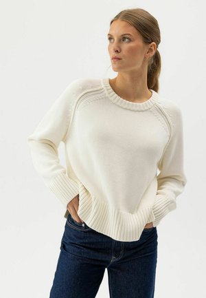 Jauna sieviete ar gaiši brūniem matiem zirgastē, ģērbusies krēmkrāsas adītā džemperī un tumši zilās džinsās, stāv ar rokām kabatās, skatoties sānis.