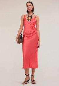 Rochie din satin coral cu un design drapat, decolteu în formă de V și lungime până la jumătatea gambelor, asociată cu tocuri negre cu siret și o geantă țesută.