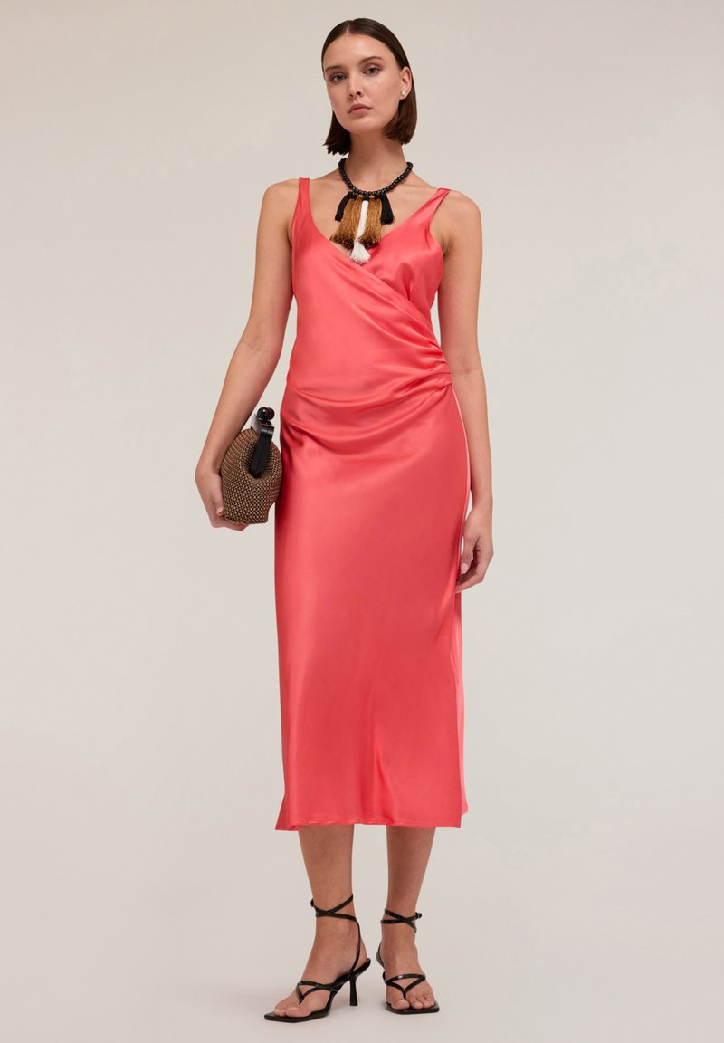 Rochie din satin coral cu un design drapat, decolteu în formă de V și lungime până la jumătatea gambelor, asociată cu tocuri negre cu siret și o geantă țesută.