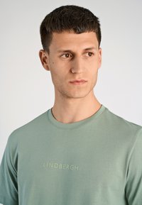 Młody mężczyzna o krótkich ciemnych włosach, ubrany w zieloną koszulkę z napisem „LINDBERGH”, spoglądający nieco w prawo na jasnym tle.