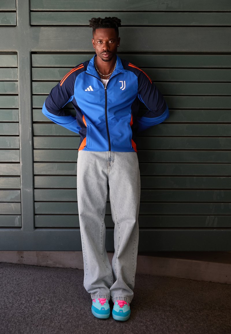En mann står mot en grønn vegg, iført en blå og oransje sportsjakke, vide jeans og fargerike sneakers med rosa detaljer.