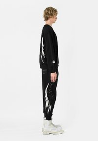 Sudadera negra con diseño gráfico de alas blancas en la parte posterior, combinada con pantalones joggers negros que presentan un patrón similar. Botas blancas.
