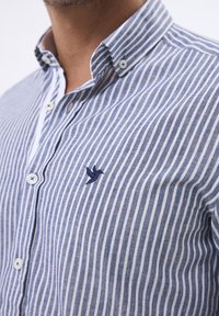 Camisa abotonada con rayas verticales en azul y blanco, cuello corto, logo de ave bordado y botones blancos. Tejido texturizado.