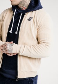 Giacca bomber in corduroy beige con texture a coste e un maglione blu con cappuccio sotto. Include cordini bianchi e dettagli con logo ricamato.