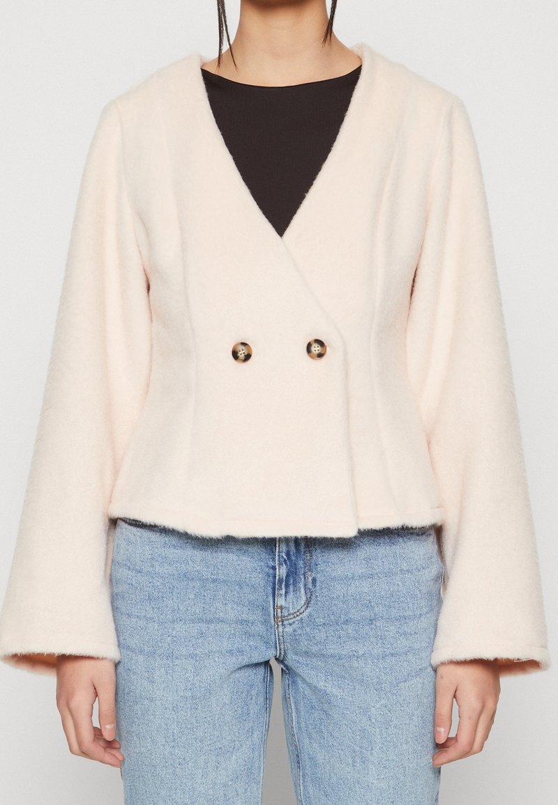 Femme portant une veste duveteuse rose clair à manches larges, boutons doubles en écaille, un haut noir en dessous et un jean bleu clair.