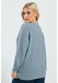 Kobieta z blond włosami, ubrana w jasnoniebieski sweter o grubym splocie, spogląda przez ramię na tle jednolitego jasnego tła.