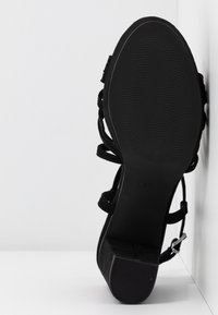 Sandale à talons en daim noir avec une semelle texturée, présentant des brides entrecroisées et un accent de boucle en métal. Marquage de taille visible au bas.