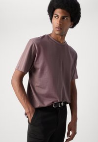 Tricou cu mânecă scurtă și guler rotund, în nuanțe de mov pastelat, realizat dintr-un material moale, purtat cu pantaloni negri și o curea alb-negru cu dungi.
