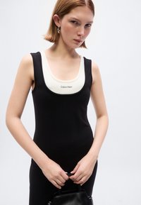 Μαύρο αμάνικο φόρεμα με κρεμ μπούστο σε σχήμα scoop, με λογότυπο Calvin Klein. Το μοντέλο κρατά μια μικρή μαύρη τσάντα χειρός. Ομαλή υφή.