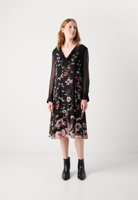 Schwarz-blumiges Kleid mit durchsichtigen langen Ärmeln, das mit pinken und grünen Designs verziert ist. Der Ausschnitt hat einen rüschigen Akzent und wird mit schwarzen Knöchelschuhen kombiniert.