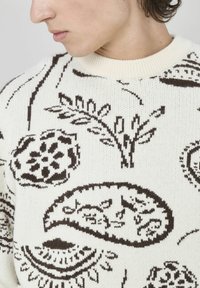 Pull en tricot crème avec des motifs botaniques marron foncé, comprenant des fleurs et des feuilles, avec un col rond côtelé et un tissu texturé.