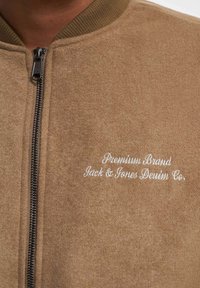 Brun zip-up jakke med teksturert finish, ribbet krage og hvit brodert merkevare på venstre side. Sølvfarget glidelåsdetaljer synlige.